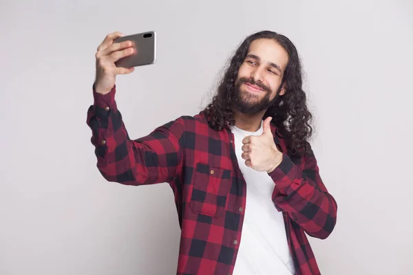 年轻的成人男子在红色格子衬衫和黑色长卷发站立和使自拍与智能手机和显示拇指在灰色背景 — 图库照片