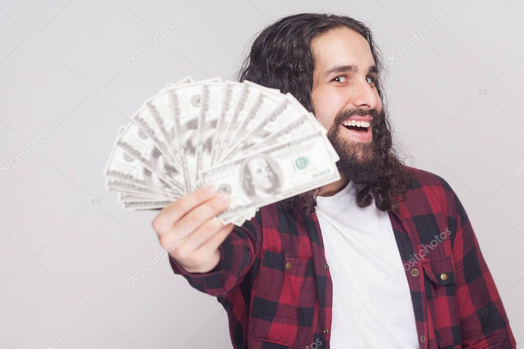Happy kindness young adult businessman in red checkered shirt and long curly hair holding dollars and looking at camera with open mouth on grey background