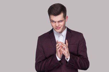 Portrait of funny cunning handsome young man in violet suit and white shirt looking at camera with cheating face on grey background clipart