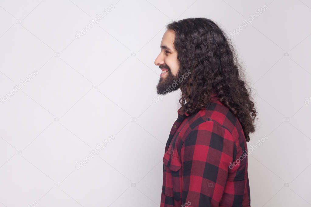 side view portrait of handsome happy man with beard and black long curly hair in checkered red shirt standing with toothy smile on grey background.