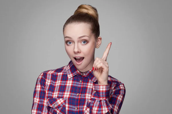 Satisfied beautiful woman in pink checkered shirt with collected bun hairstyle looking at camera with toothy smile and pointing finger up on gray background