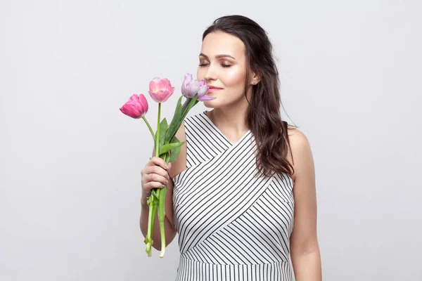 Portrait of young brunette beautiful woman with makeup in white striped dress holding tulips and smelling with closed eyes on grey background.