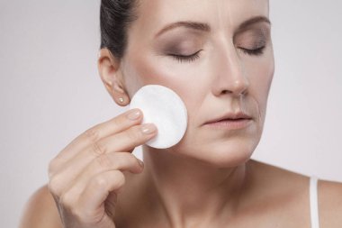 Olgun kadın yüzünden gri arka plan üzerinde gelen makyaj kaldırmak için micellar su ile pamuk panelini kullanma