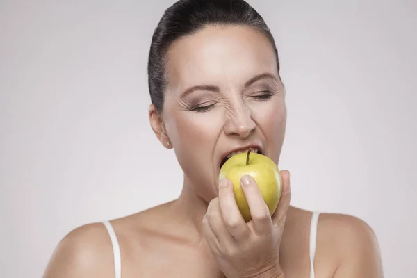 Portrait of middle aged woman eating yellow apple on grey background