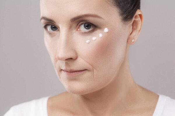 Middle aged woman applying cream on skin near eyes on grey background