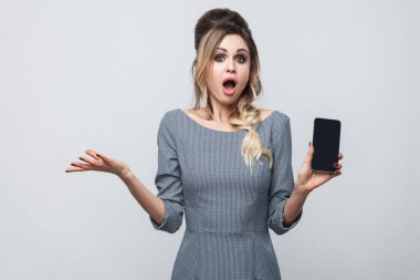 Akıllı telefon tutarak ve hayret suratla kameraya bakarak gri elbiseli şok güzel modern kadın portresi 