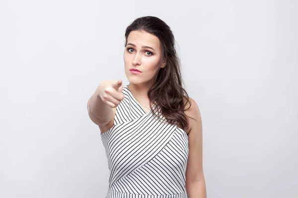 You. Portrait of serious beautiful young brunette woman with makeup and striped dress looking and pointing at camera on grey background.