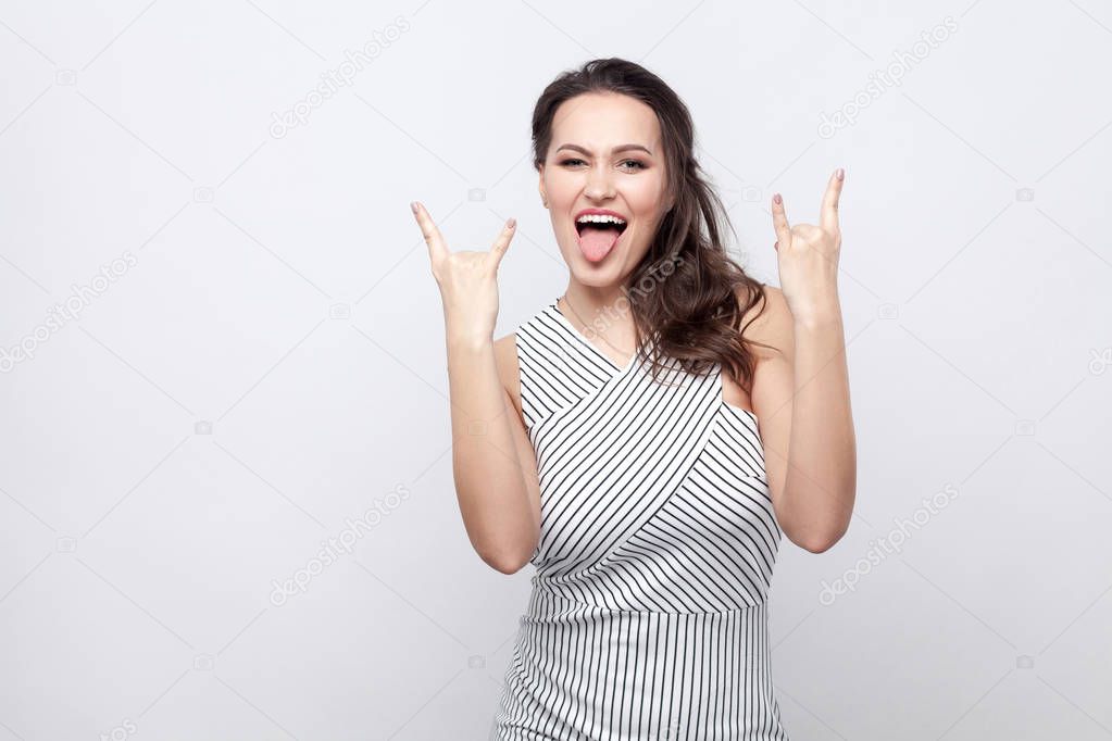 Portrait of fuuny crazy beautiful young brunette woman with makeup and striped dress standing and looking at camera with rock signs and tongue out on grey background
