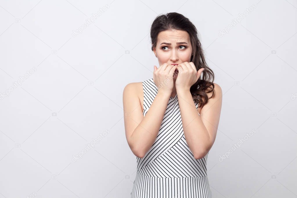 Portrait of worry beautiful young brunette woman with striped dress looking away and biting nails with stressed face on grey background.