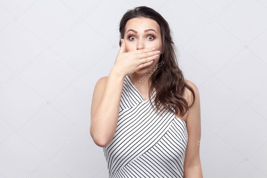 I will be silence. Portrait of shocked beautiful young brunette woman with makeup and striped dress standing with big eyes and covering her mouth on grey background.