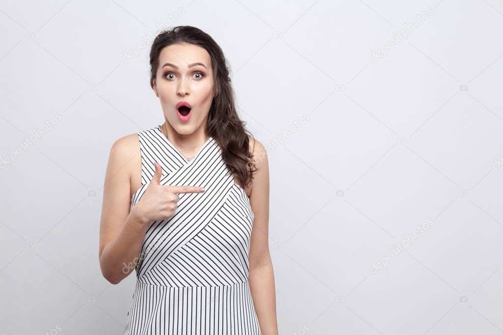 Portrait of beautiful amazed young brunette woman in striped dress standing and looking at camera with surprised face and pointing aside on grey background