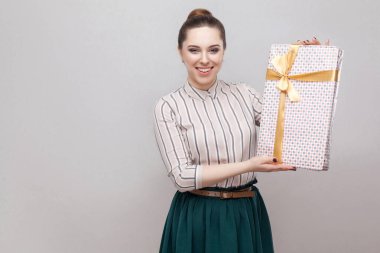 Çizgili gömlekli ve sarı yay ile mevcut tutarak ve kameraya gri arka plan üzerinde seyir yeşil etek iyilik çekici genç kadın