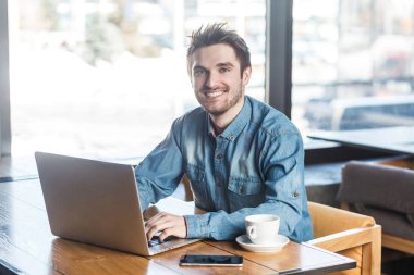 Kafede oturan ve laptop fotoğraf makinesi ararken dişlek gülümseme ile mesaj yazarak kot gömlek yakışıklı neşeli sakallı genç işadamı portresi 