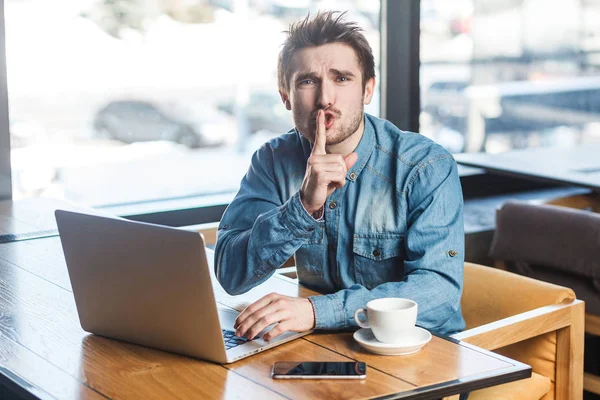 Portrait of handsome severe bearded freelancer in blue jeans shirt leaning finger on mouth and showing silence sign at camera while sitting with laptop in cafe