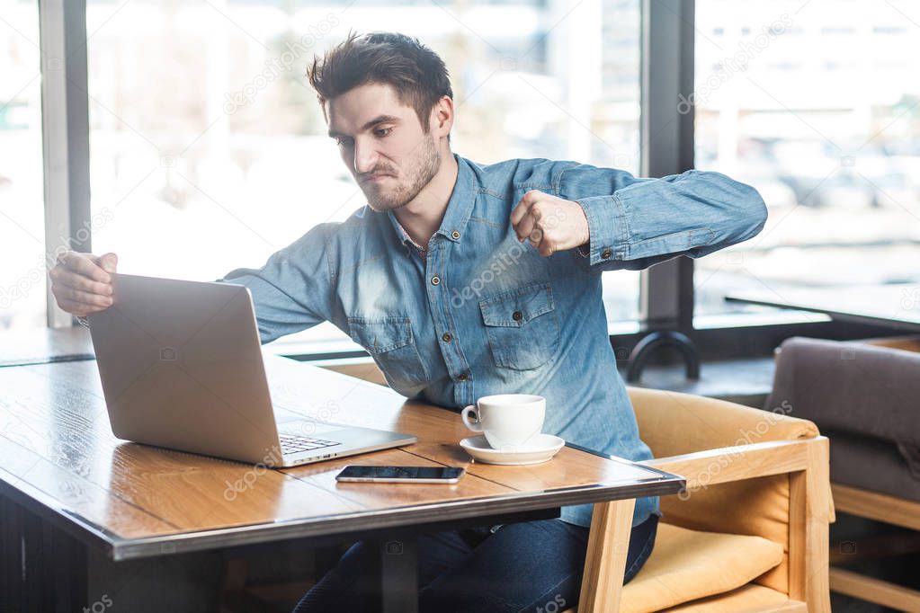 portrait of aggressive unhappy businessman in blue jeans shirt sitting in cafe and having bad mood while ready to punch worker through webcam with raised fist 
