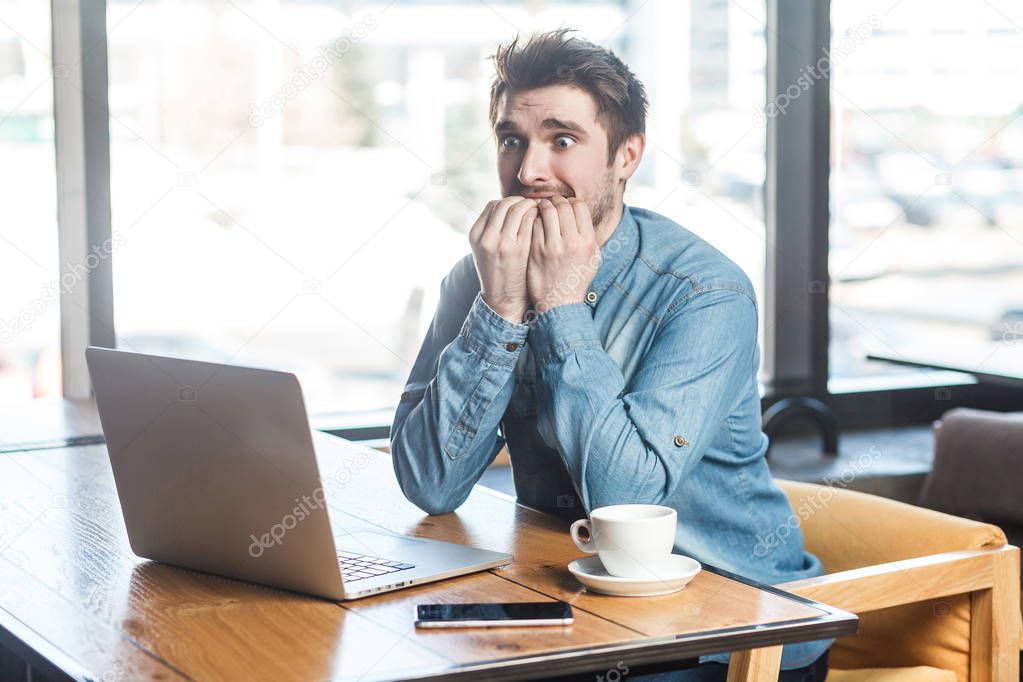 portrait of emotional nervous businessman in blue jeans shirt sitting in cafe and biting nails while looking at laptop 