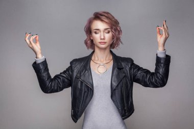 Portrait of calm beautiful woman with short pink hairstyle and makeup in black leather jacket standing in meditation yoga pose with raised arms and closed eyes   clipart