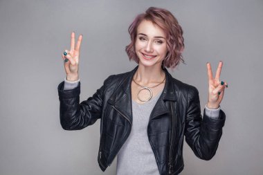Mutlu dişlek gülen kısa pembe saç ve makyaj zafer işaretleri ile ayakta ve kameraya seyir siyah deri ceketli güzel kadınla 