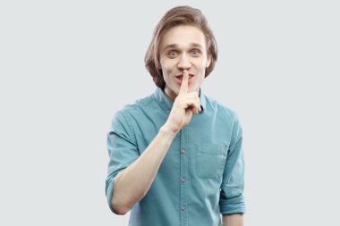 Mavi gömlekli kameraya sessizlik işaretiyle ışık gri arka plan üzerinde arıyorsunuz komik uzun saçlı sarışın adam portresi.