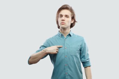Portrait of proud long haired blonde man in blue shirt standing and pointing himself while looking at camera on light grey background. clipart