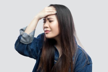 Endişeli güzel esmer Asyalı genç kadın açık gri renkli kafasına üzerinde elini tutarak rahat mavi kot ceketli portresi.