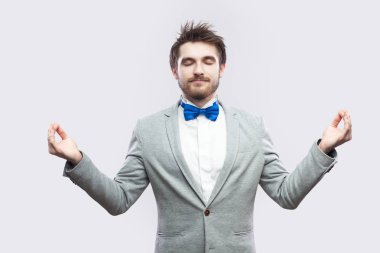 sakin sakin ol rahat gri elbiseli sakallı yakışıklı ve mavi papyon ile ayakta kollarını kaldırdı ve yoga yaparken ve ışık gri arka plan üzerinde meditasyon sırasında gözleri kapalı