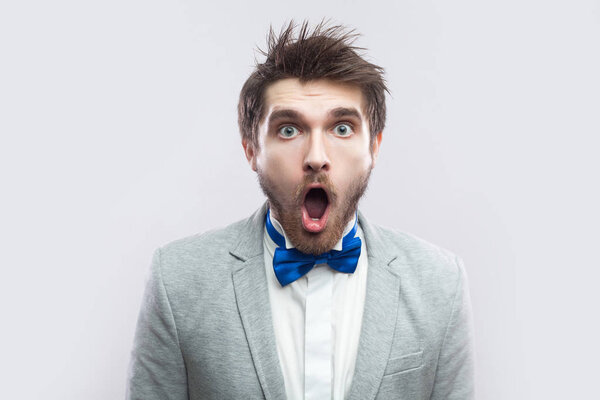 shocked handsome bearded man in casual grey suit and blue bow tie standing with big eyes and open mouth and looking at camera on light grey background.