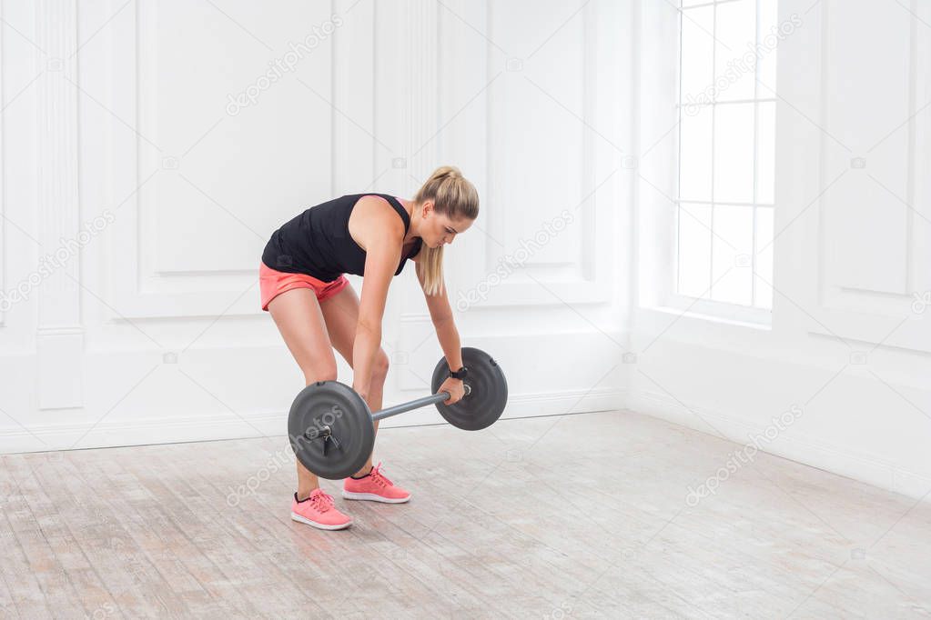 Side view of athletic blonde woman in pink shorts and black top doing wrong squats with barbell at gym 