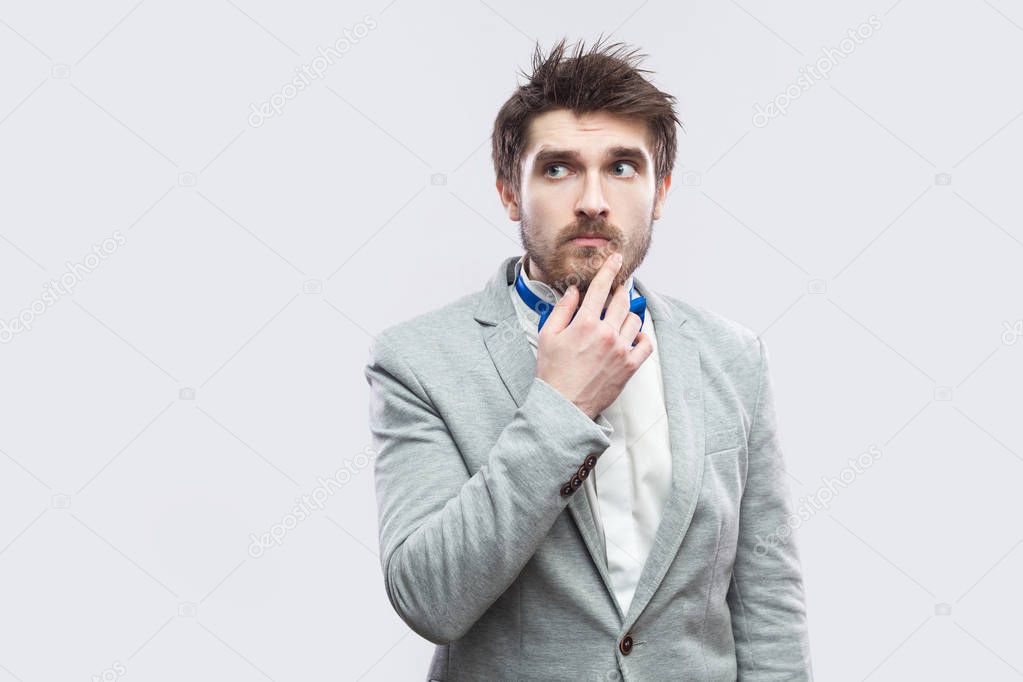 doubtful handsome bearded man in casual grey suit and blue bow tie touching his chin and looking away while thinking on light grey background.