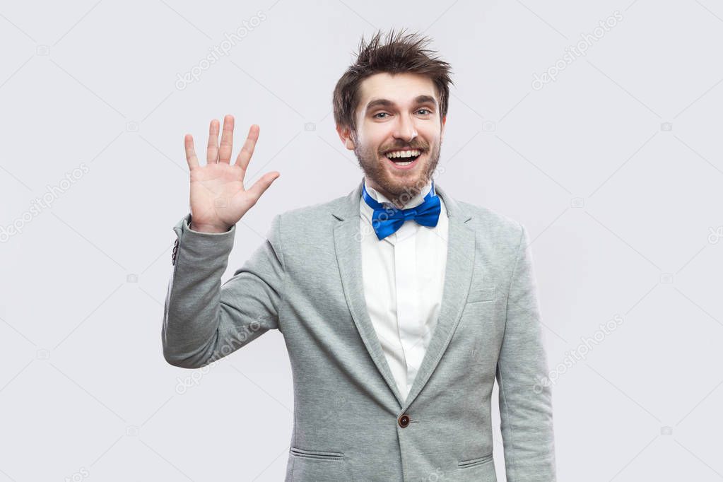 excited happy handsome bearded man in casual grey suit and blue bow tie standing and looking at camera, greeting and toothy smile. indoor studio shot, isolated on light grey background