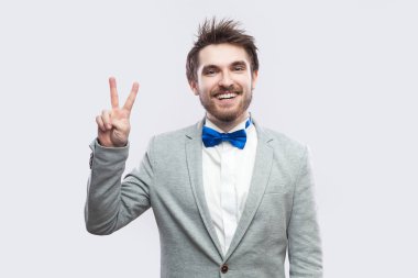 mutlu dişlek gülen yakışıklı adam rahat gri elbiseli sakallı ve mavi papyon ayakta ve zafer ile kameraya bakıyor ışık gri arka plan üzerinde oturum açın.
