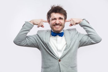 sıradan gri renk takımlı ve mavi ile kapalı gözler ayakta ve parmakların kulaklarında ışık gri arka plan üzerine koyarak papyon endişeli sakallı yakışıklı.