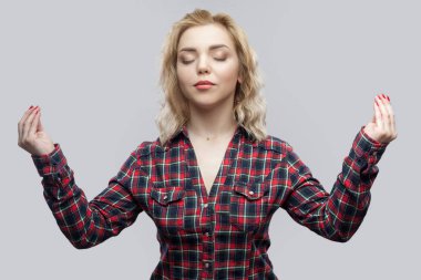 calm beautiful blonde young woman in casual red checkered shirt standing with raised arms and closed eyes while meditating on grey background. clipart