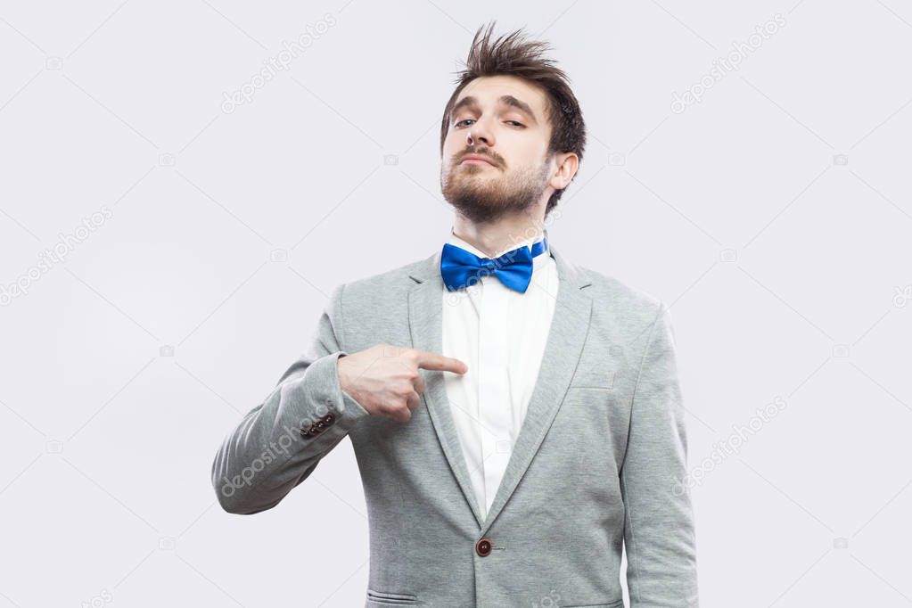 proud satisfied haughty handsome bearded man in casual grey suit and blue bow tie pointing himself and looking at camera on light grey background.