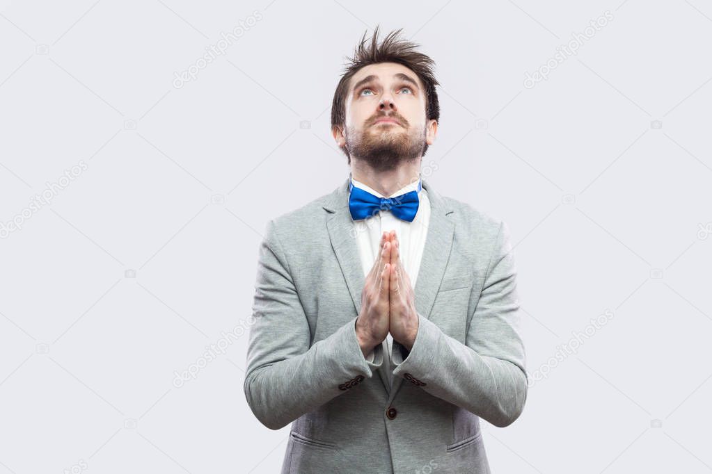 Worried handsome bearded man in casual grey suit and blue bow tie standing with palm hands and begging god to help on light grey background.