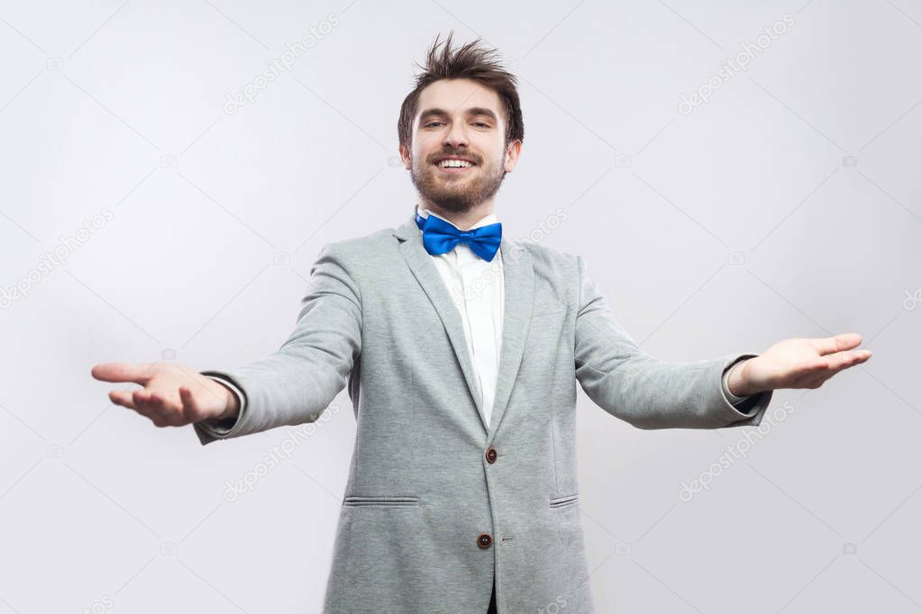 happy handsome bearded man in casual grey suit and blue bow tie standing with divorced arms and looking at camera with toothy smile on light grey background.