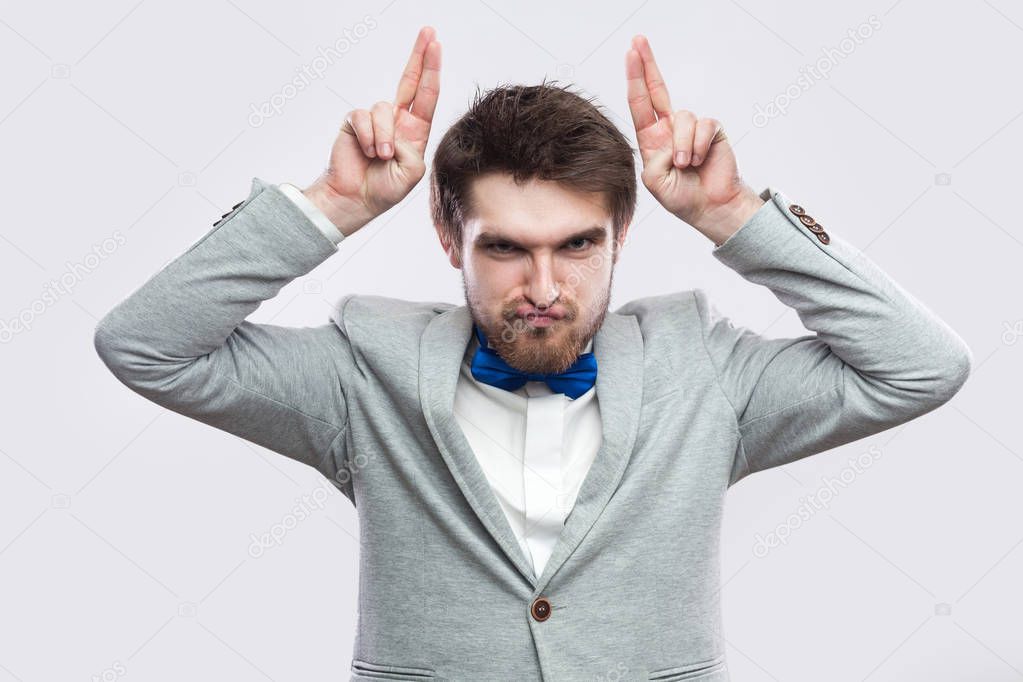 playful handsome bearded man in casual grey suit and blue bow tie looking at camera with funny face and showing horns finger on light grey background.