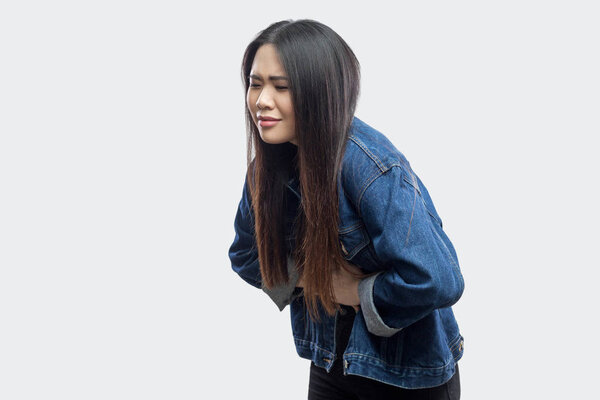 Side view of beautiful brunette asian young woman in casual blue denim jacket feeling pain on her stomach on light grey background.