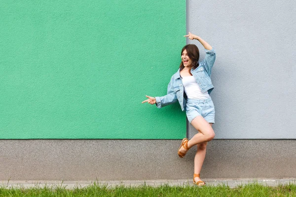 快乐满意美丽的女人在休闲牛仔裤牛仔风格站立和指向一边与两只手接近绿色和蓝色墙壁在夏天 — 图库照片