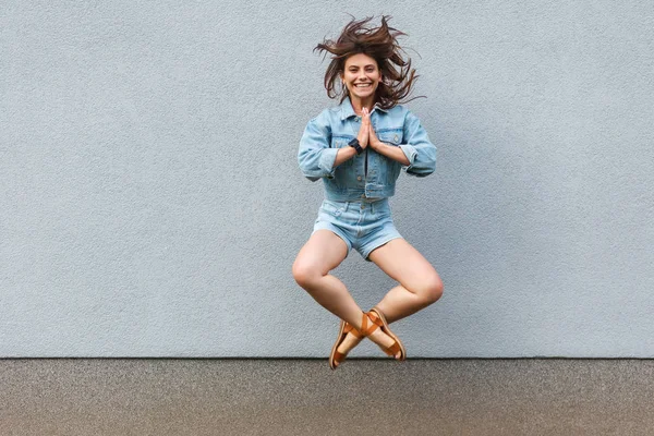 Free happy beautiful woman in casual jeans denim style jumping with namaste palm hands at light blue wall and looking at camera with toothy smile, happiness concept