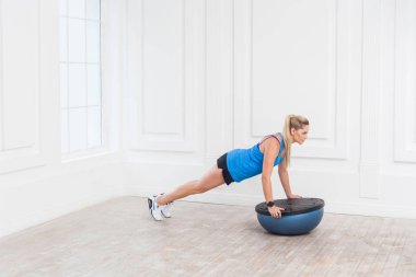 Sportif güzel genç atletik sarışın kadın siyah şort ve mavi en iyi spor salonunda bosu denge eğitmen olarak karın kasları için tahta yaparken çalışma odaklı 