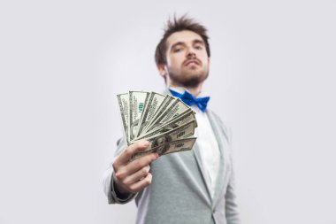 confident young businessman in gray coat and blue bow tie standing proudly with head up and giving dollars on gray background. clipart