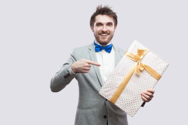Gri arka plan üzerinde kameraya ararken sarı yay ile günümüze kadar memnun genç adam klasik gri ceket ve mavi ayakta ve parmak işaret papyon