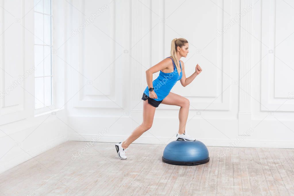 Side view of sporty blonde woman in black shorts and blue top making one step on bosu balance while working in gym 