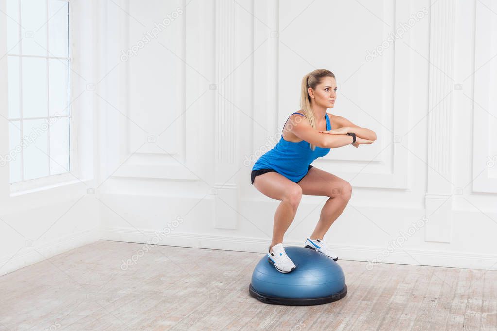 sporty blonde woman in black shorts and blue top working in gym doing squat while holding balance on bosu balance trainer