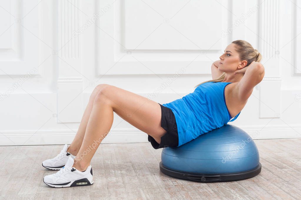 Focused sporty beautiful young athletic blonde woman in black shorts and blue top working in gym doing exercises for abdominal muscles on bosu balance trainer  