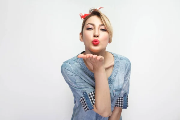 beautiful young woman in casual blue denim shirt with makeup and red headband looking and sending kiss at camera isolated on white background.