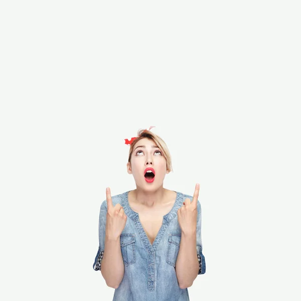 excited of young woman in casual blue denim shirt with makeup and red headband standing looking and pointing up isolated on white background.