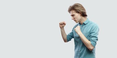 ciddi uzun saçlı sarışın yakışıklı genç boks hareketi ile ayakta ve ışık gri arka plan üzerine saldırmaya hazır mavi gömlekli yan görünüm