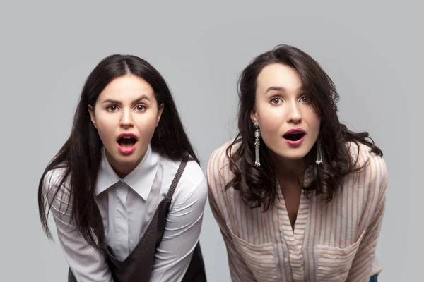 shocked beautiful brunette women in casual style standing together and looking at camera with opened mouths and big eyes on grey background